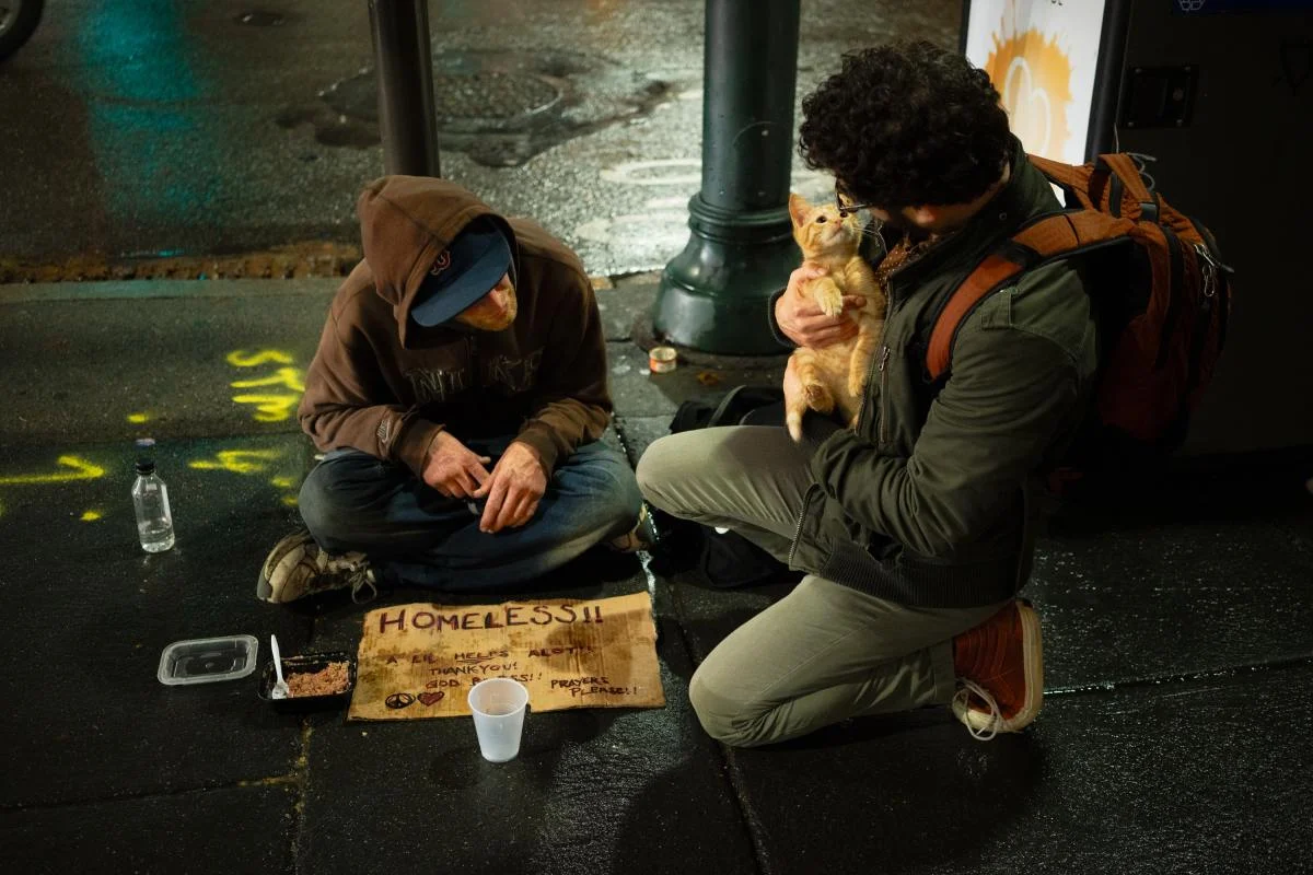 A person sharing food with someone seated on a city sidewalk